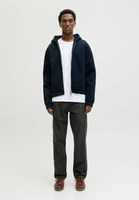 Marinblå hoodie med dragkedja och en känguruficka, kombinerad med en vit T-shirt. Grå byxor och bruna snörskor fullbordar outfiten.