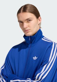 Modrá síťovaná bunda na zip s bílými pruhy na rukávech a malým bílým logem. Vyrobena s vysokým límcem a volným střihem.