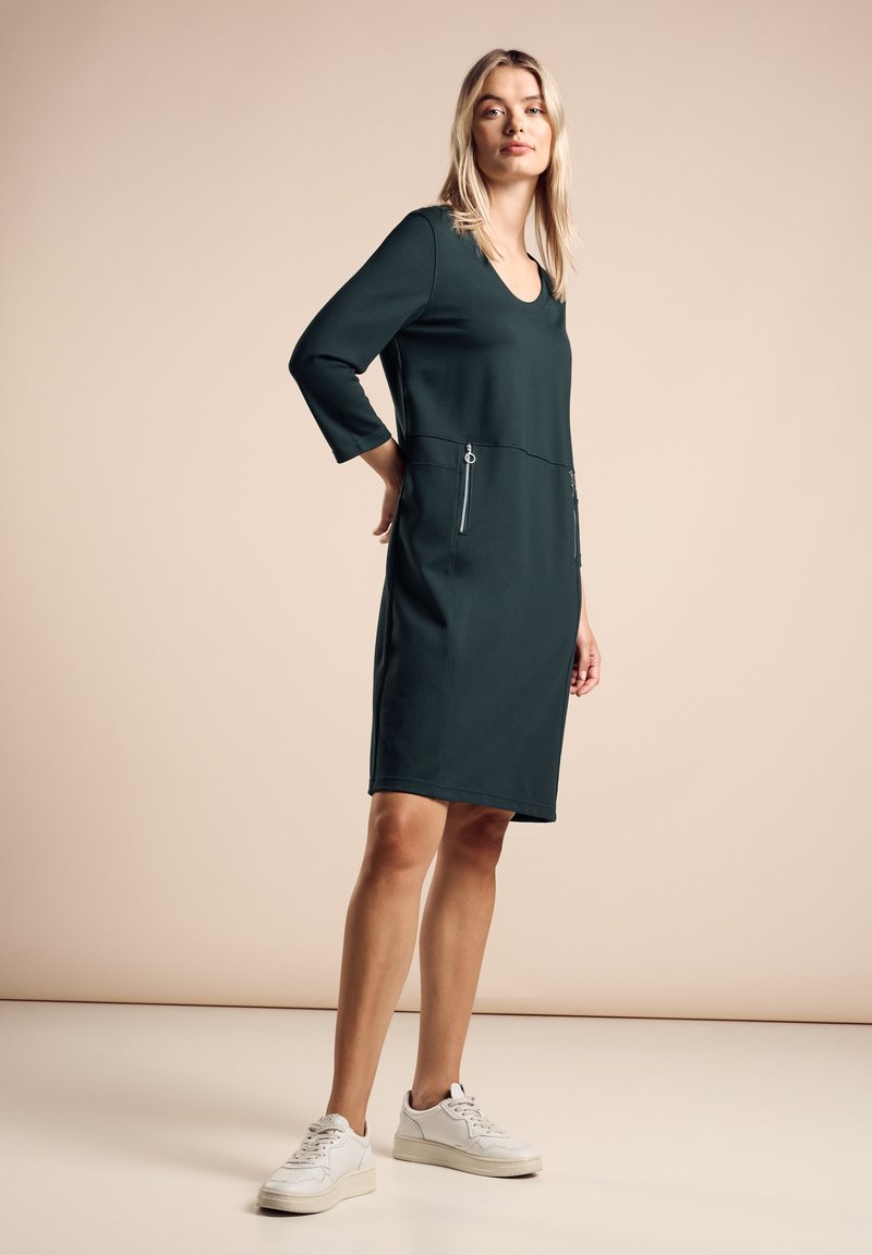 Robe verte foncée avec un col rond et des manches trois-quarts, dotée de deux poches zippées à la taille et d'une coupe au genou.