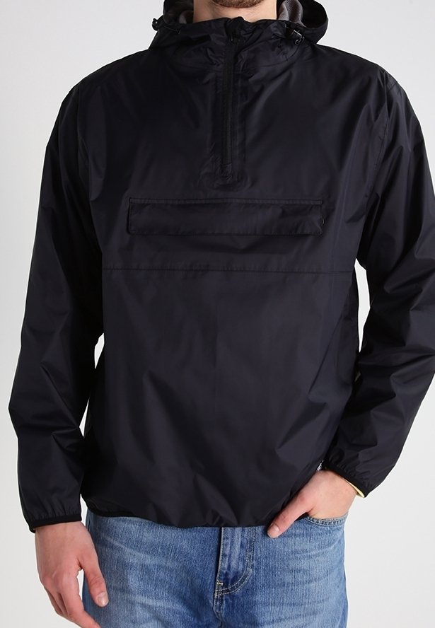 Veste coupe-vent noire à capuche avec fermeture éclair frontale et poche à rabat, portée par une personne avec la main dans la poche d’un jean bleu.