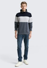 Gestreifter Sweatshirt in Grau und Marineblau mit hohem Kragen, kombiniert mit blauen Jeans und weißen Sneakern. Der Sweatshirt hat einen strukturierten Stoff.