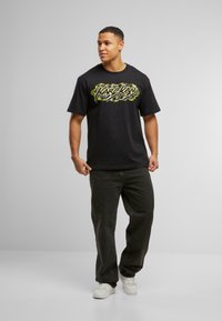 Camiseta de algodón negra con un diseño gráfico amarillo en la parte delantera. Combinada con jeans oscuros y zapatillas blancas. Corte estándar, mangas cortas.