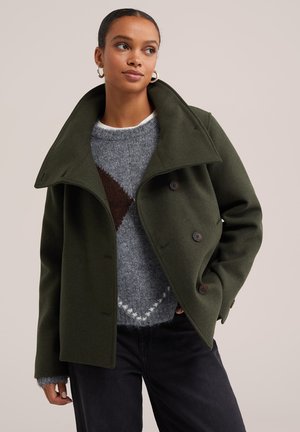 Giovane donna che indossa un cappotto verde oliva doppiopetto sopra a un maglione grigio con fantasia e pantaloni neri, guarda di lato con la mano in tasca.