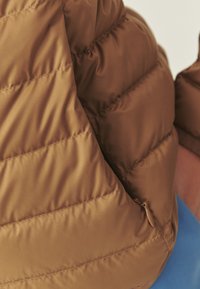Veste matelassée marron avec une poche zippée partiellement visible, portée par une personne vêtue de bleu.