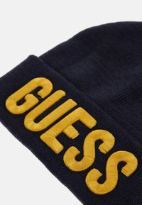 Granatowa dziana czapka z wyraźnym żółtym haftem z napisem "GUESS". Miękka w dotyku z podwiniętym brzegiem.