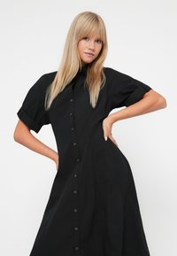 Donna bionda in un vestito nero con bottoni e maniche corte arrotolate posa con le mani sui fianchi contro uno sfondo bianco semplice.