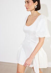 Valge puff-sleeve kleit ruudukujulise dekoltee, ülikitsas keha ja laieneva seeliku ning sileda kangaga, millel on kerge sära ja pehme viimistlus.