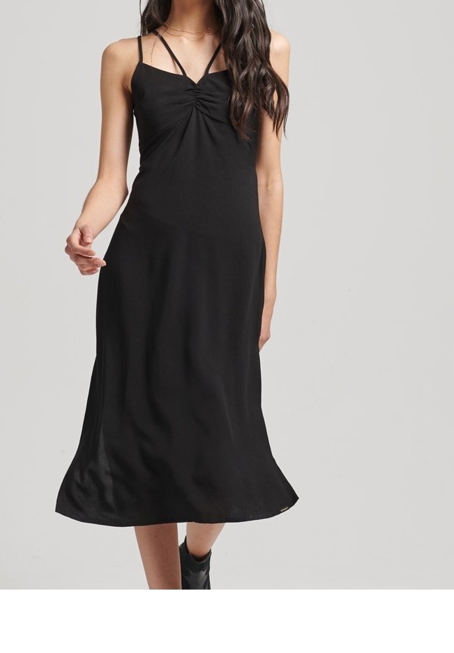 Femme portant une robe midi noire à fines bretelles et détail froncé, associée à des bottines noires, debout devant un fond clair.