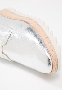 Chaussure métallique argentée avec une finition texturée, un accent en liège sur la semelle, et une base en caoutchouc blanc avec un motif en zigzag.