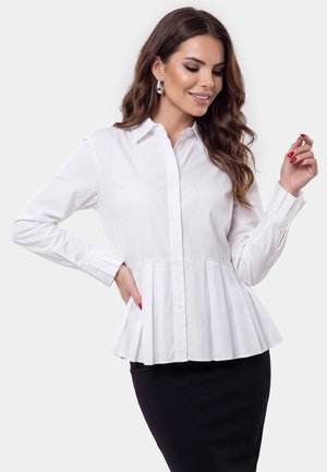 Vrouw met lang bruin haar die een witte geplooide blouse met knopen en een zwarte rok draagt, glimlacht en poseert met één hand op haar heup.