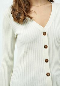 Kermanau ribbed cardigan dengan lengan panjang, leher v, dan lima kancing coklat yang melintang di bagian depan. Tekstur lembut dengan desain pas.
