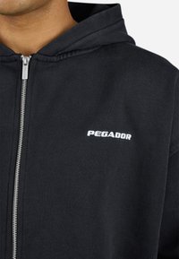 Black zip-up hoodie made of cotton blend. Features a cozy hood, metal zipper, and white "PEGADOR" logo on the chest. Smooth texture.
