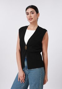 Μαύρο αμάνικο ζακέτα με V-λαιμόκοψη, χρυσό κούμπωμα μπροστά, υφασμάτινη υφή, σε συνδυασμό με ανοιχτόχρωμα τζιν ψηλόμεσα παντελόνια.