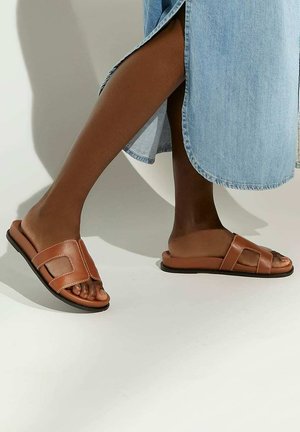 Brown leather slide sandals with two wide straps and visible stitching, paired with a light denim skirt. Flat black sole, open-toe design.