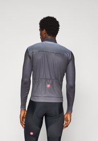 Mann i grå langermet sykkeltrøye og svarte polstrede sykkelshorts, står vendt bort fra kamera mot en ensfarget bakgrunn.
