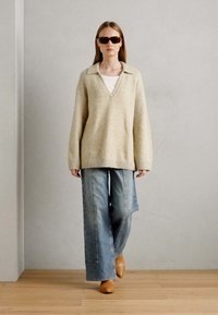 Beige strikket oversized trøje med V-udskæring, parret med lyseblå denim jeans med brede ben og brune lædermules; minimalistisk stil.