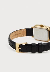 Bracelet en cuir noir avec une boucle et un accent en métal doré. Cadran de montre carré visible, présentant un fond argenté avec le logo de la marque embossé.