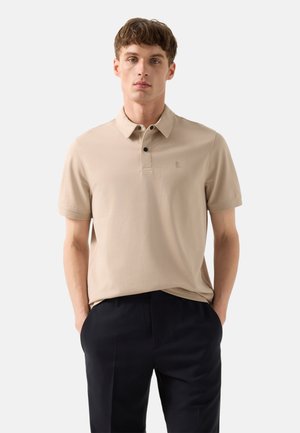 Młody mężczyzna ubrany w beżową koszulkę polo z krótkim rękawem i czarne spodnie, stoi z rękami w kieszeniach na jednolitym tle.