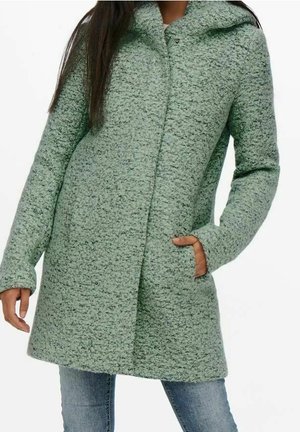 Femme portant un manteau à capuche texturé vert clair avec manches longues et poches latérales, associé à un jean bleu.