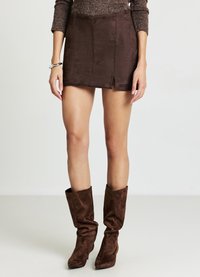 Brauner Minirock aus Wildleder mit einem vorderen Schlitz und glatter Textur. Kombiniert mit kniehohen braunen, lässigen Stiefeln.