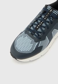 Αθλητικό παπούτσι με μπλε και μαύρο μοτίβο, σκούρες μπλε κορδόνια, λευκή σόλα και κείμενο «J.LINDEBERG» στην γλώσσα του παπουτσιού.