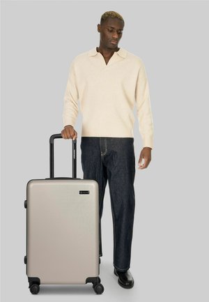 Uomo con maglione beige e jeans scuri che tiene la maniglia di una valigia rigida argento con quattro ruote su sfondo grigio.