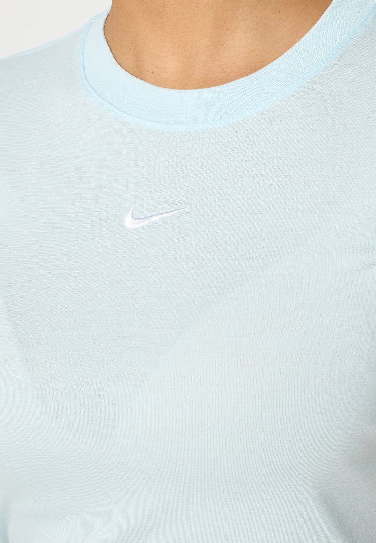 Jasnoniebieska koszulka z okrągłym dekoltem z małym białym logo Nike w formie haftu na klatce piersiowej, noszona przez osobę.
