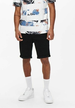 Mann trägt ein weißes Hemd mit tropischem Muster, schwarze hochgekrempelte Shorts, weiße Sportsocken und weiße Sneakers, steht vor einem schlichten Hintergrund.