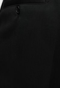 Κοντινό πλάνο μαύρου υφασμάτινου παντελονιού που δείχνει το κλείσιμο με κουμπιά στο μπροστινό μέρος και μέρος μιας πλάγιας λοξής τσέπης.