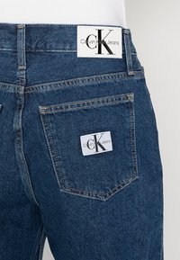 Mörkblå denimjeans med en slät textur, med en Calvin Klein Jeans-logotyp på baksidan av midjan och bakfickorna.