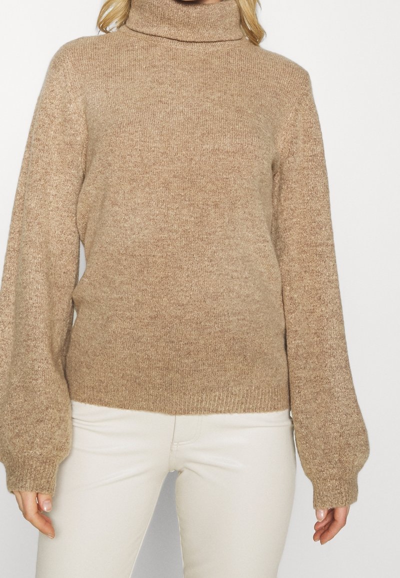 Beige coltrui met lange, volumineuze mouwen en een gestructureerd breipatroon. Ribgebreide boorden en zoom voegen detail toe. G gedragen met lichtgekleurde broek.