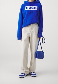 Modrá mikina s bílým logem "HUGO", béžové široké kalhoty, modrá crossbody taška a modro-bílé tenisky. Hladký materiál a pohodlný design.