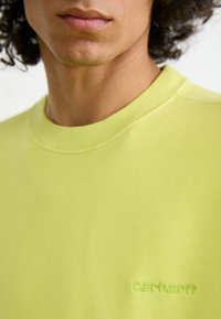 Lys limegrøn sweatshirt med rund hals. Blødt stof og synligt Carhartt-logo broderet i matchende grøn. Enkel design og tekstur.