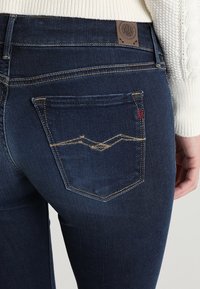 Jeans de mezclilla oscura con una textura suave, que presentan costuras amarillas contrastantes, una parche de cuero en la cintura y un diseño distintivo en el bolsillo trasero.