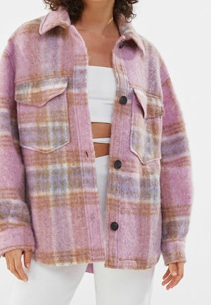 Femme portant une veste en laine à carreaux surdimensionnée rose, marron et bleue avec de grands boutons noirs, par-dessus un top court blanc et un pantalon blanc.