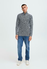 Pull à col zippé quart gris et blanc marbré avec un design de tricot texturé. Porté avec un jean bleu clair et des baskets blanches.