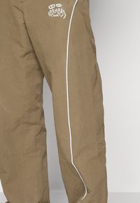 Calças cáqui folgadas com uma costura branca nas laterais, apresentando um logótipo gráfico impresso em branco na zona superior da coxa.