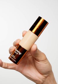 Podkład w postaci serum w miękkim, beżowym płynie umieszczony w cylindrycznej butelce z błyszcząca różowo-złotą zakrętką i szczegółowym brandingiem.
