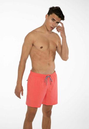Korallfarbene Badeshorts mit einem Kordelzugbund, kontrastierenden schwarzen und weißen Bindebändern sowie seitlichen Taschen. Das Material wirkt leicht und glatt.