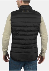 Schwarwest gepolsterte Weste mit vertikalen Nähten, hohem Kragen und Druckknopfverschluss hinten, getragen über einem beigen Langarmshirt.