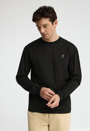 Hombre con camisa negra de manga larga con pequeño logo blanco en el pecho y pantalones beige, ajustándose las mangas, sobre un fondo liso y claro.