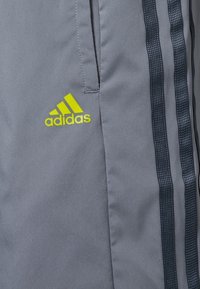 Šedé sportovní kalhoty Adidas s černými a žlutými akcenty. Mezi rysy patří ikonický design se třemi pruhy a hladký, lehký materiál.