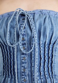 Top de denim sem alças com textura franzida, apresentando um laço nas costas, botões à frente e tonalidades de azul.
