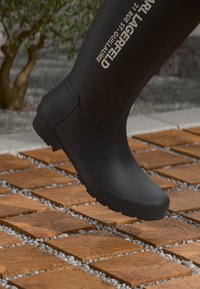 Črne gumijaste škornje z gladko površino, ki imajo teksturiran podplat, izbočeno logotipno oznako in zaobljeno konico, postavljene nad ploščicami.