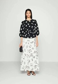 Blusa preta com flores e mangas bufantes, combinada com uma saia branca fluida com estampa floral preta. Modelo segura uma pequena bolsa preta.