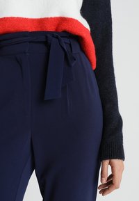 Marineblauwe hoge taillebroek met een strikriem, gecombineerd met een gebreide trui met een wit lijfje en een rode zoom. Zachte textuur, casual ontwerp.