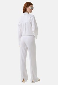 Witte sportoutfit met een kraag, lange mouwen en een rugplissé, vergezeld van bijpassende broek met gespleten zomen. Gelaagde stof, relaxte pasvorm.