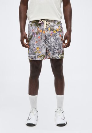Mann trägt weiße Sneakers, weiße Socken, weißes T-Shirt und graue Shorts mit Blumenmuster und Kordelzug, steht vor einfarbigem Hintergrund.