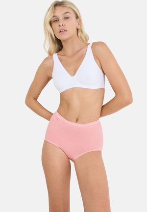 Frau mit blonden Haaren, die einen weißen BH und hoch taillierte rosa Unterwäsche trägt, steht mit einer Hand in der Hüfte vor einem schlichten Hintergrund.