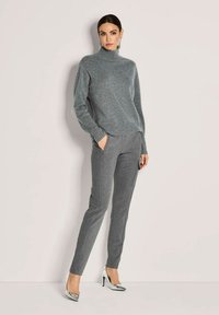 Eine Frau, die einen grauen Rollkragenpullover, graue maßgeschneiderte Hosen und silberne High Heels trägt, steht vor einem hellen Hintergrund.